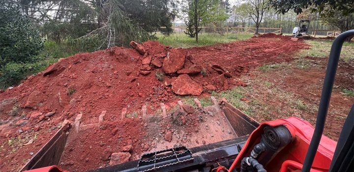 A red skid steer loader is seen working on a site with piles of red soil and rocks, surrounded by trees and fencing.