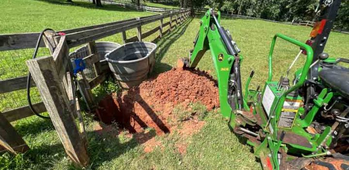A green excavator is digging a hole in red soil near a wooden fence and grassy field.