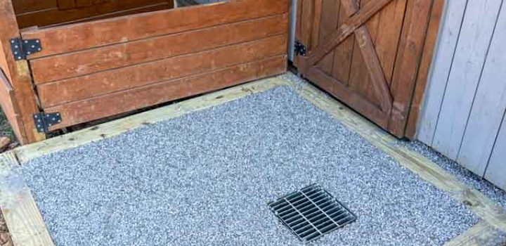 The entrance to a barn featuring a wooden door, a drainage grate in the gravel floor, and a well-maintained area surrounded by wooden fencing.