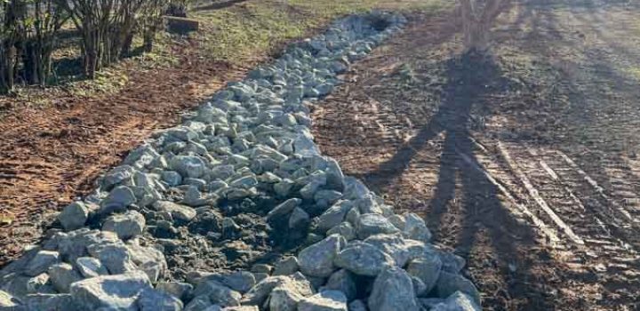 A pathway made of neatly arranged rocks runs alongside a trench, likely for drainage, with trees and a house visible in the background under bright sunlight.