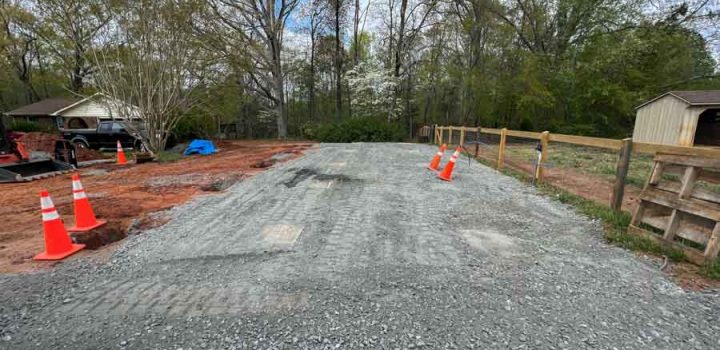 A gravel driveway under construction, marked with orange traffic cones for safety, bordered by a wooden fence and trees.