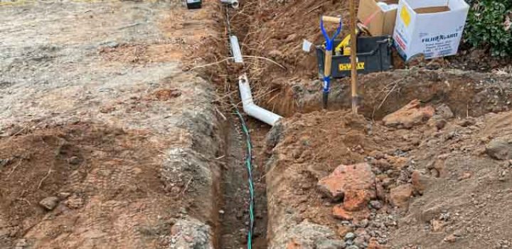 An open trench in the ground with pipes being installed, surrounded by construction materials and tools.