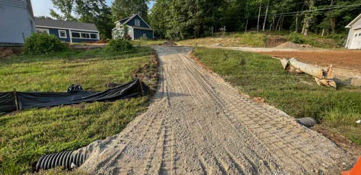 A gravel path leading towards a construction site with houses in the background, surrounded by greenery and fencing.