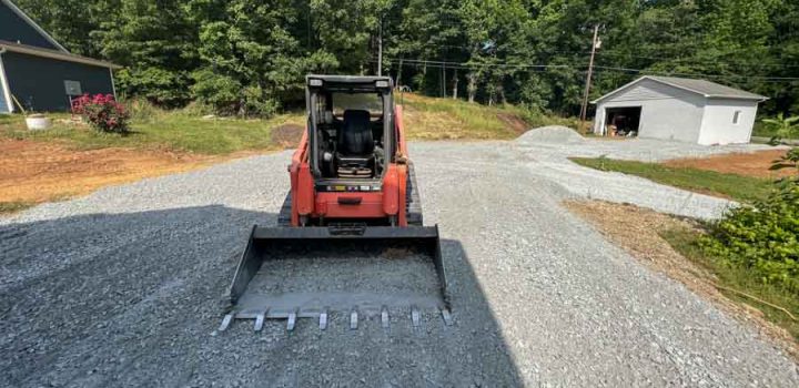 A red skid steer loader is parked on a gravel path, with a house and trees visible in the background.