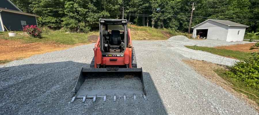 A red skid steer loader is parked on a gravel path, with a house and trees visible in the background.
